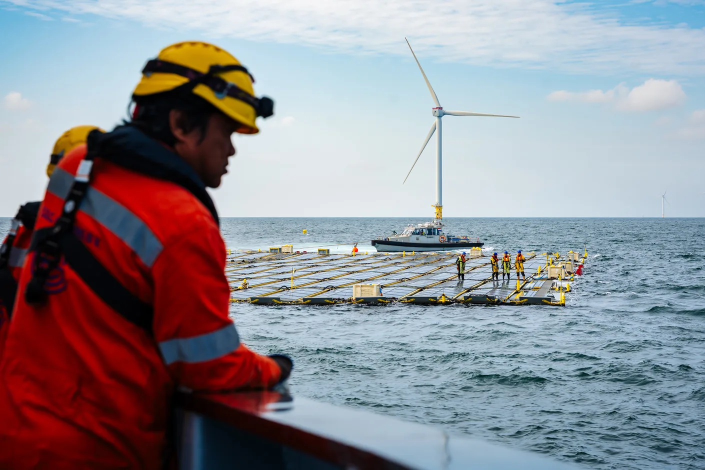 Crew member in a red offshore suit looking out over the floating solar farm with a wind turbine behind it