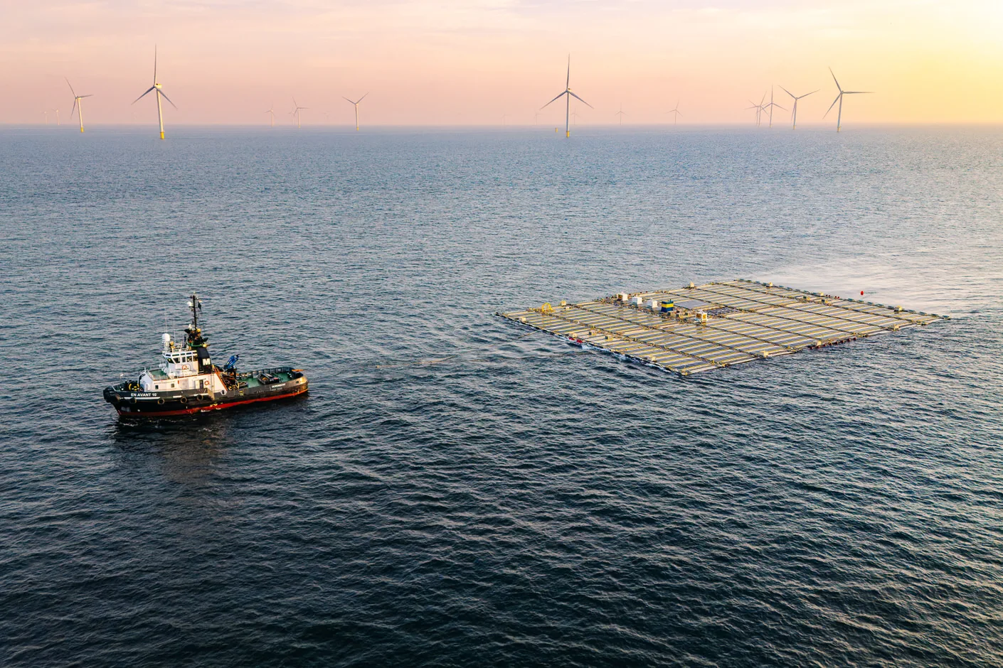 A tug pulls the floating solar farm across the sea at sunset, with wind turbines on the horizon